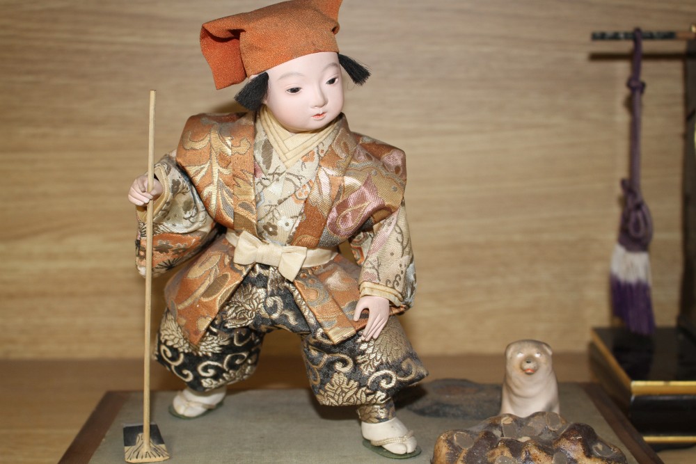 A Japanese late Meiji period composition and fabric figure of a boy farmer holding an adze with puppy seated nearby, mounted on display